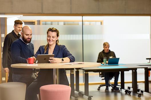 On the left in the picture are two people in the foreground at a high table, leaning over a tablet and talking. A young man is walking behind them. On the right, an older man can be seen at a desk in the background.